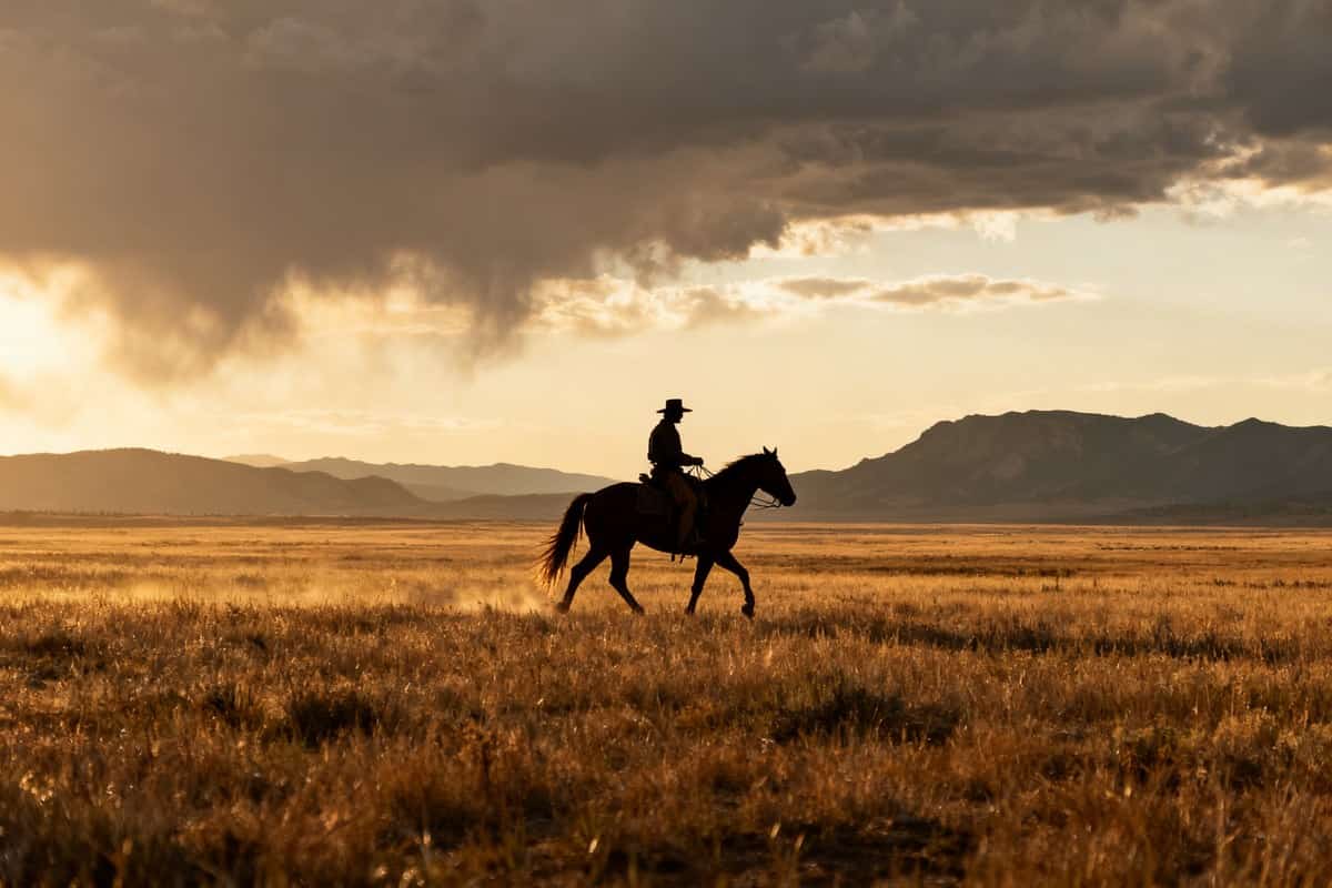 Cavalier solitaire traversant une prairie au coucher du soleil, montagnes en toile de fond, ambiance western de 1899