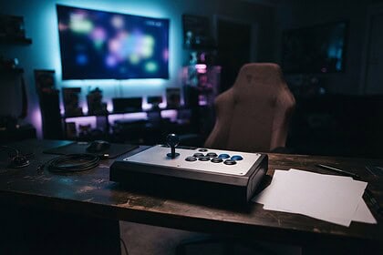Stick d'arcade posé sur un bureau en bois dans une pièce sombre, face à un fauteuil de jeu vide et un grand écran flou