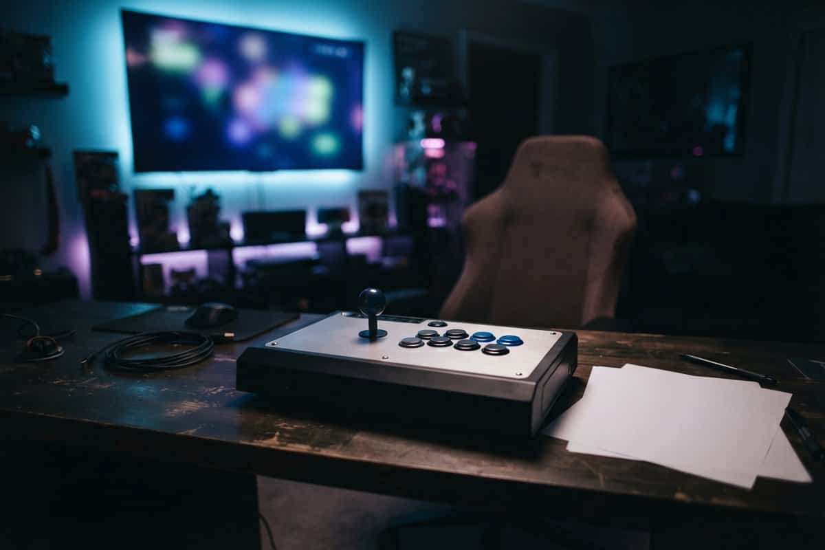 Stick d'arcade posé sur un bureau en bois dans une pièce sombre, face à un fauteuil de jeu vide et un grand écran flou