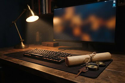 Bureau en bois éclairé par une lampe, clavier mécanique, parchemin roulé et boussole devant un écran aux couleurs floues
