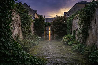 Cour pavée bordée de murs de pierre couverts de lierre, avec une porte en bois ouverte sur une lumière chaude