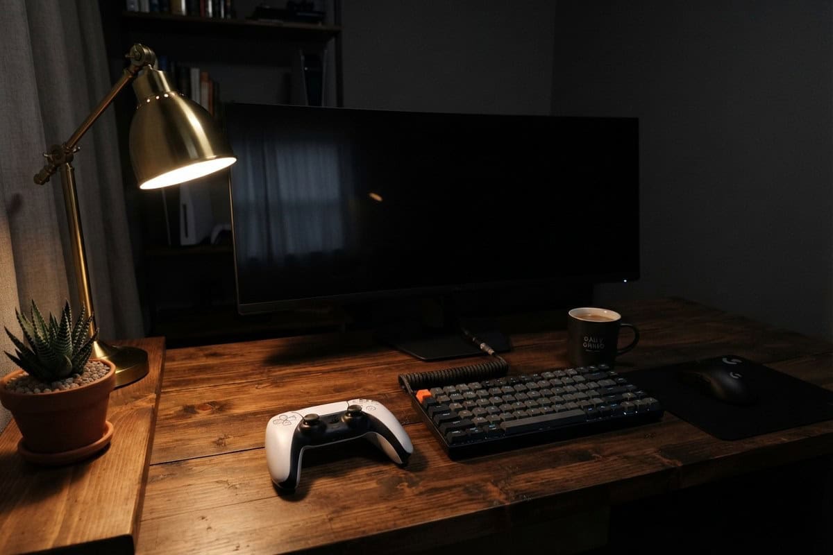 Manette blanche, clavier mécanique et mug sur un bureau en bois éclairé par une lampe, devant un écran éteint