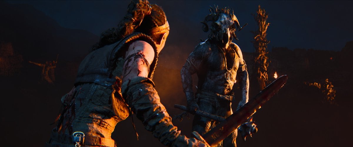 Senua, le bras ensanglanté et l'épée en main, fait face à un guerrier au masque cornu éclairé par des torches