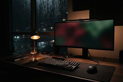 Bureau de jeu dans une pièce sombre, écran large aux couleurs floues rouges et vertes, lampe allumée et pluie sur la vitre