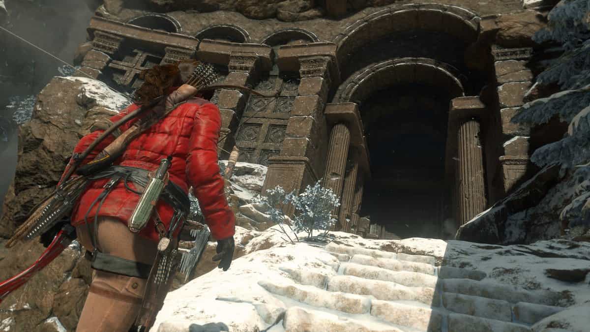 Lara Croft en parka rouge, arc dans le dos, devant l'entrée d'un tombeau creusé dans la roche enneigée